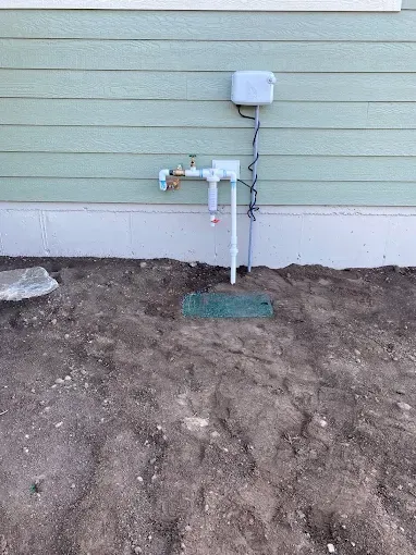An outdoor water pipe assembly with a brass spigot, a filter, and a small electrical box, set against a pale green siding.
