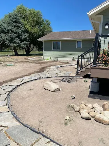 A stone pathway borders a tan yard with scattered rocks, next to a green house with a raised wooden deck.