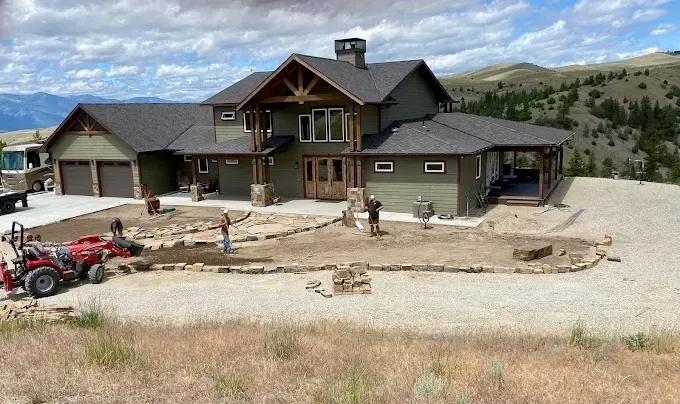Workers install stone landscaping in front of a large, olive green two-story house with a brown roof and a gravel yard.
