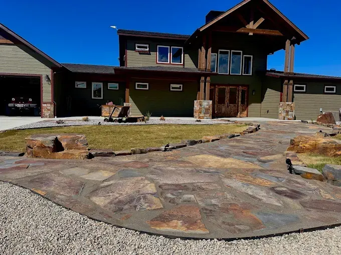 A multi-story green house with a large stone patio, a gravel driveway, and a wooden entrance under a covered porch.