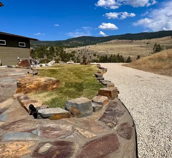 A stone patio with a rock-bordered lawn edges a gravel driveway, overlooking a dry, rolling hill landscape under blue sky.