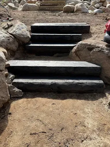 Black landscape timber steps set into a dirt hillside bordered by large decorative boulders.