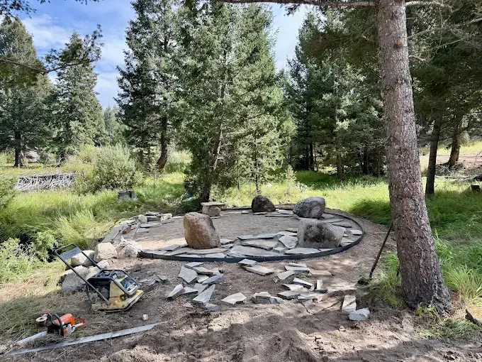 A circular stone patio under construction in a wooded area, surrounded by scattered rocks and landscaping equipment.