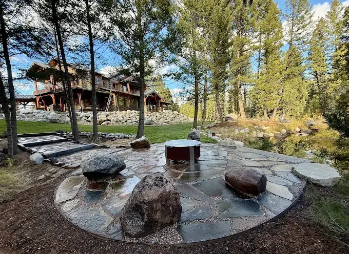 A stone patio with a fire pit and large boulders sits on a grassy lawn near a pond, overlooking a large rustic lodge home.