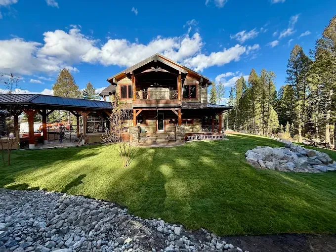 A large wooden mountain-style lodge with a covered patio, surrounded by a green lawn and trees under a blue sky.