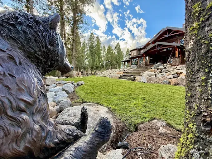 A bear sculpture sits in the foreground, looking toward a wooden lodge set among rocks and pine trees on a sunny day.