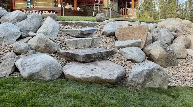 Stone steps built into a landscaped rock slope, leading up to a house with a wooden porch.