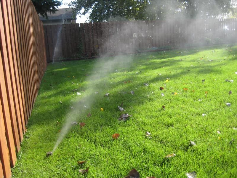 A lawn sprinkler spraying a mist of water across a green grass yard next to a wooden fence.
