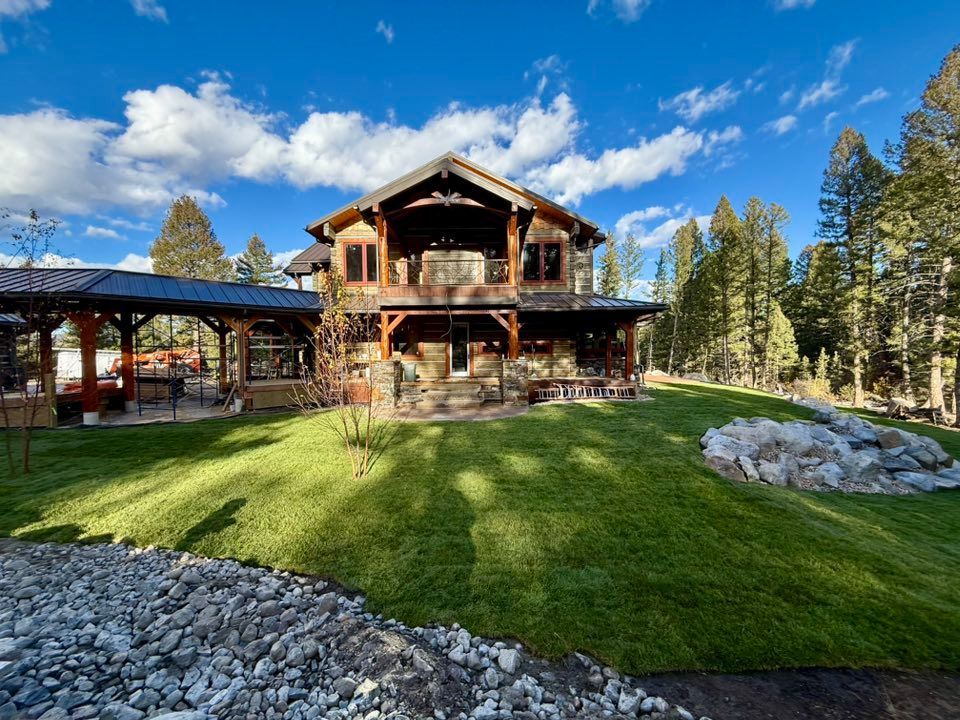 A large, rustic log cabin home with a covered patio, stone foundation, and expansive lawn under a bright blue sky.