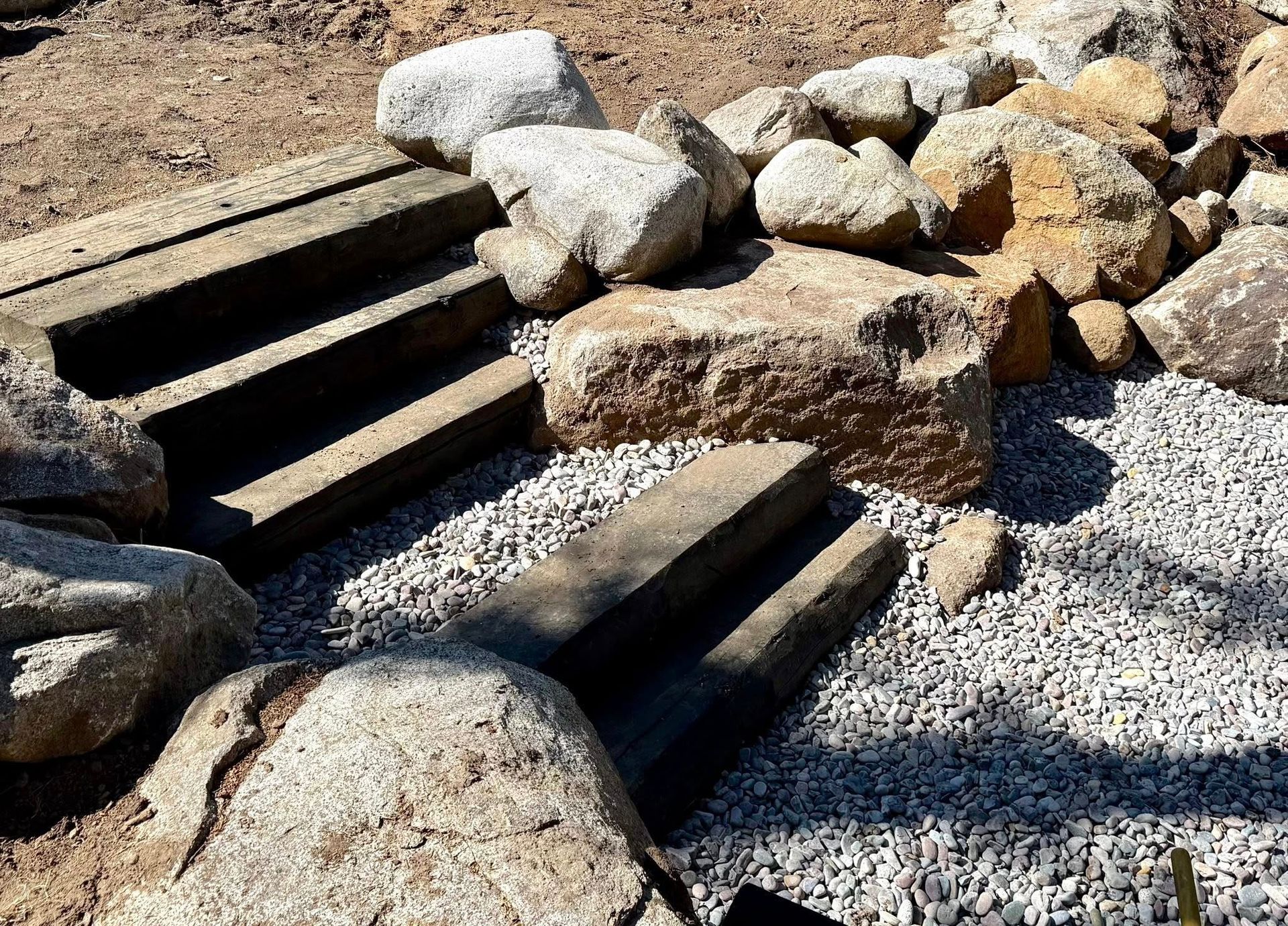 A set of weathered wooden steps built into a sloped landscape, flanked by large, textured boulders and gray gravel.