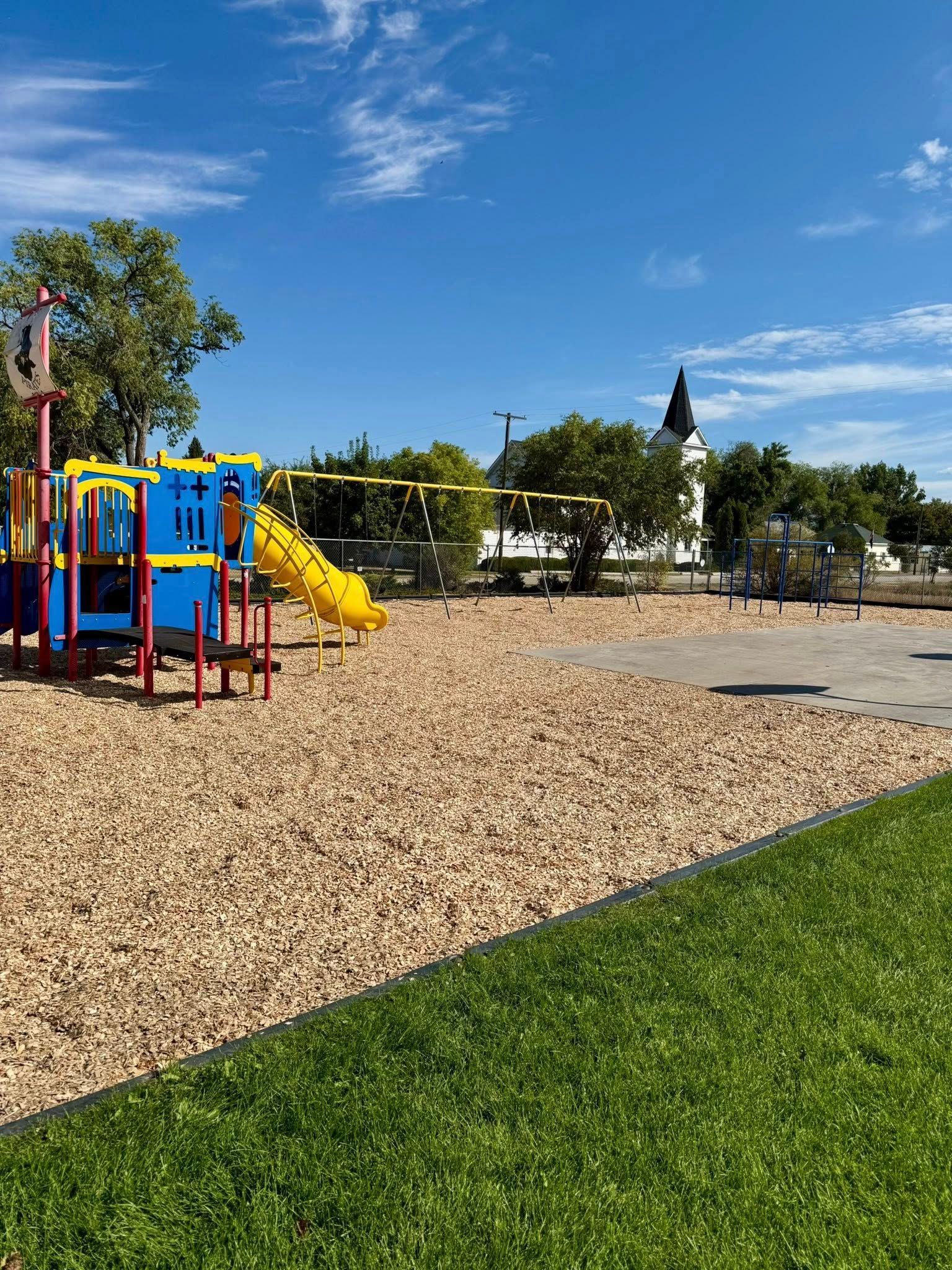 A vibrant blue and yellow playground structure sits on a wood-chip surface under a sunny sky with a church in the distance.