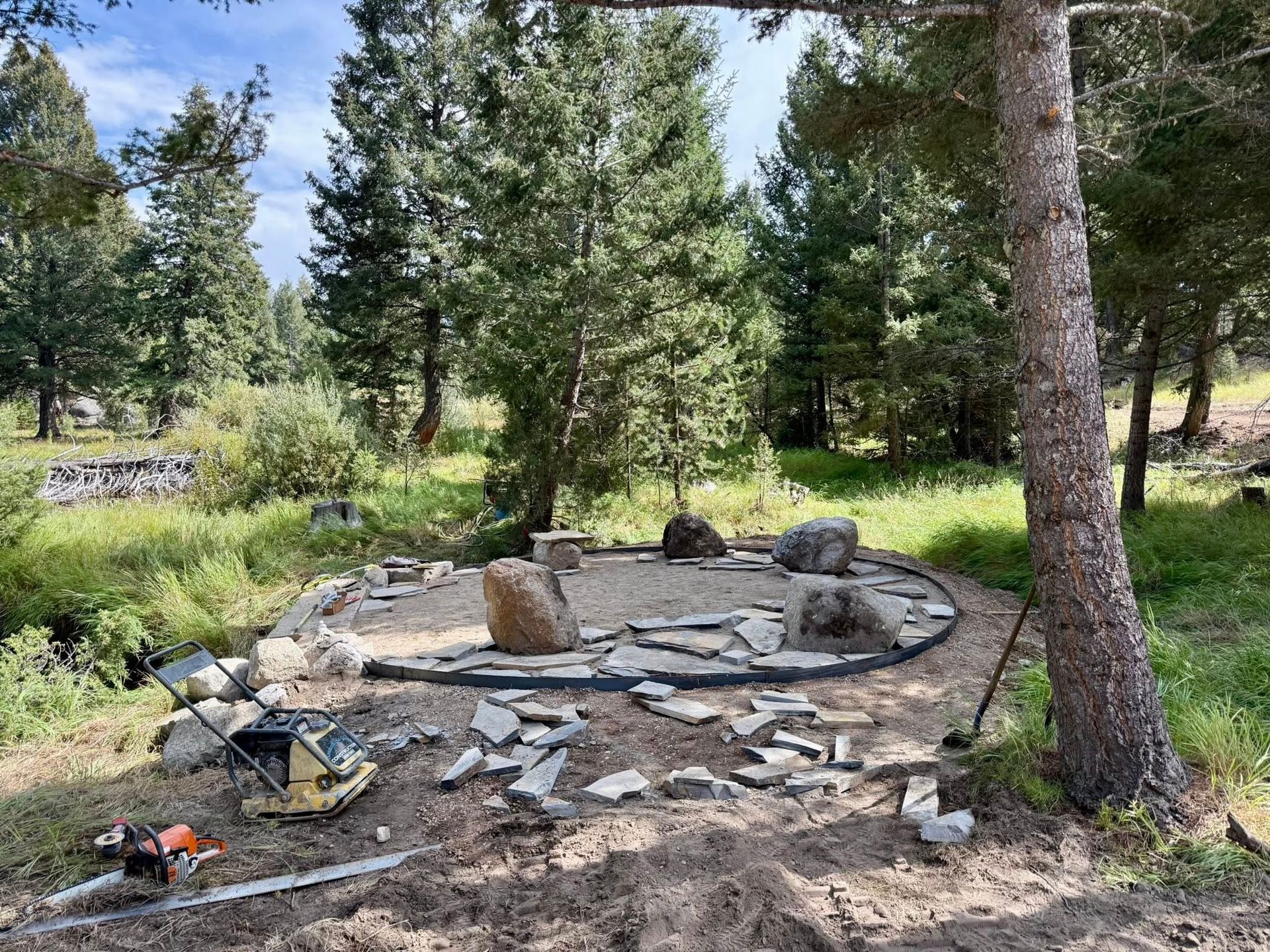 A partially constructed circular stone patio sits in a forest clearing with tools and scattered flagstones nearby.