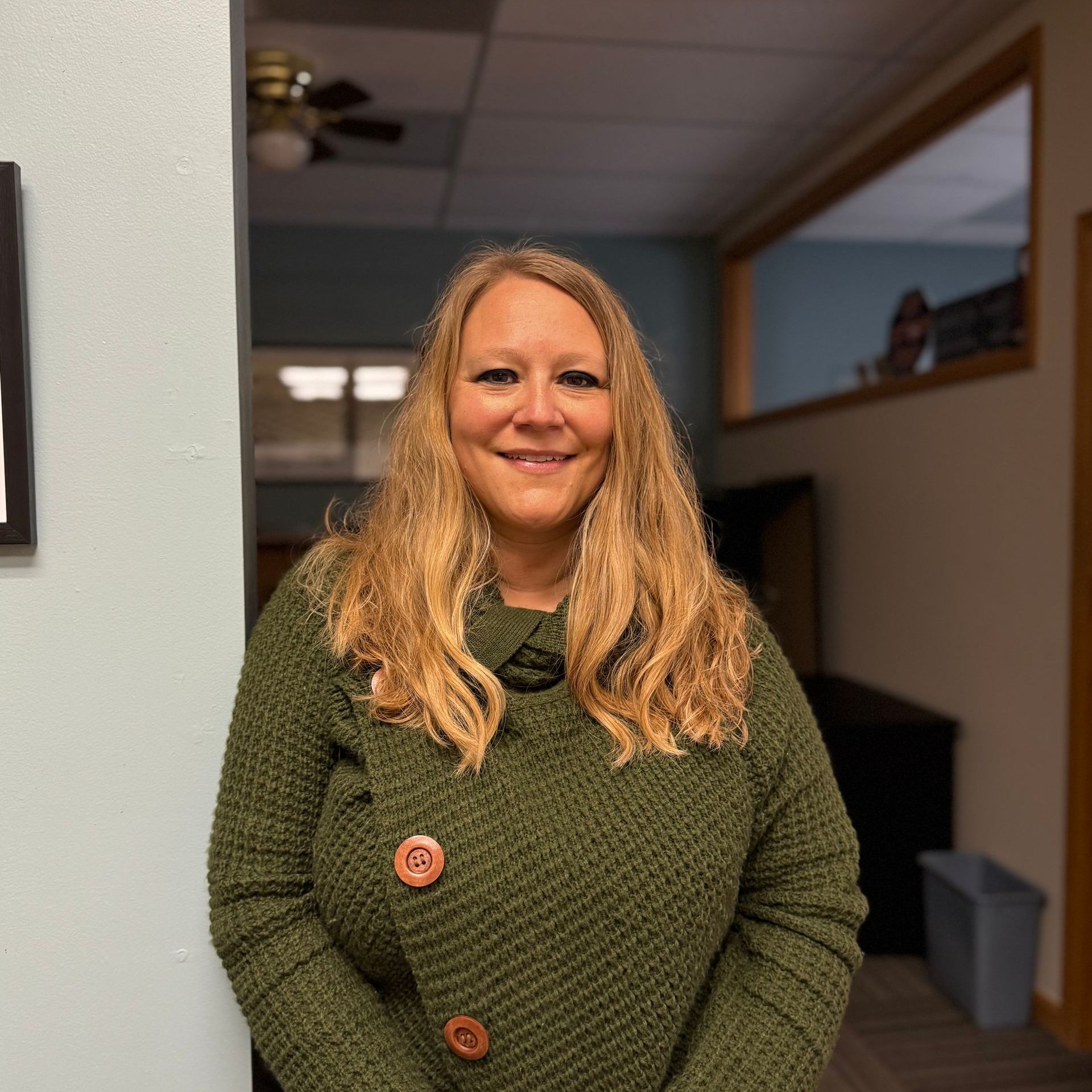 A woman in a green sweater is smiling in a hallway.