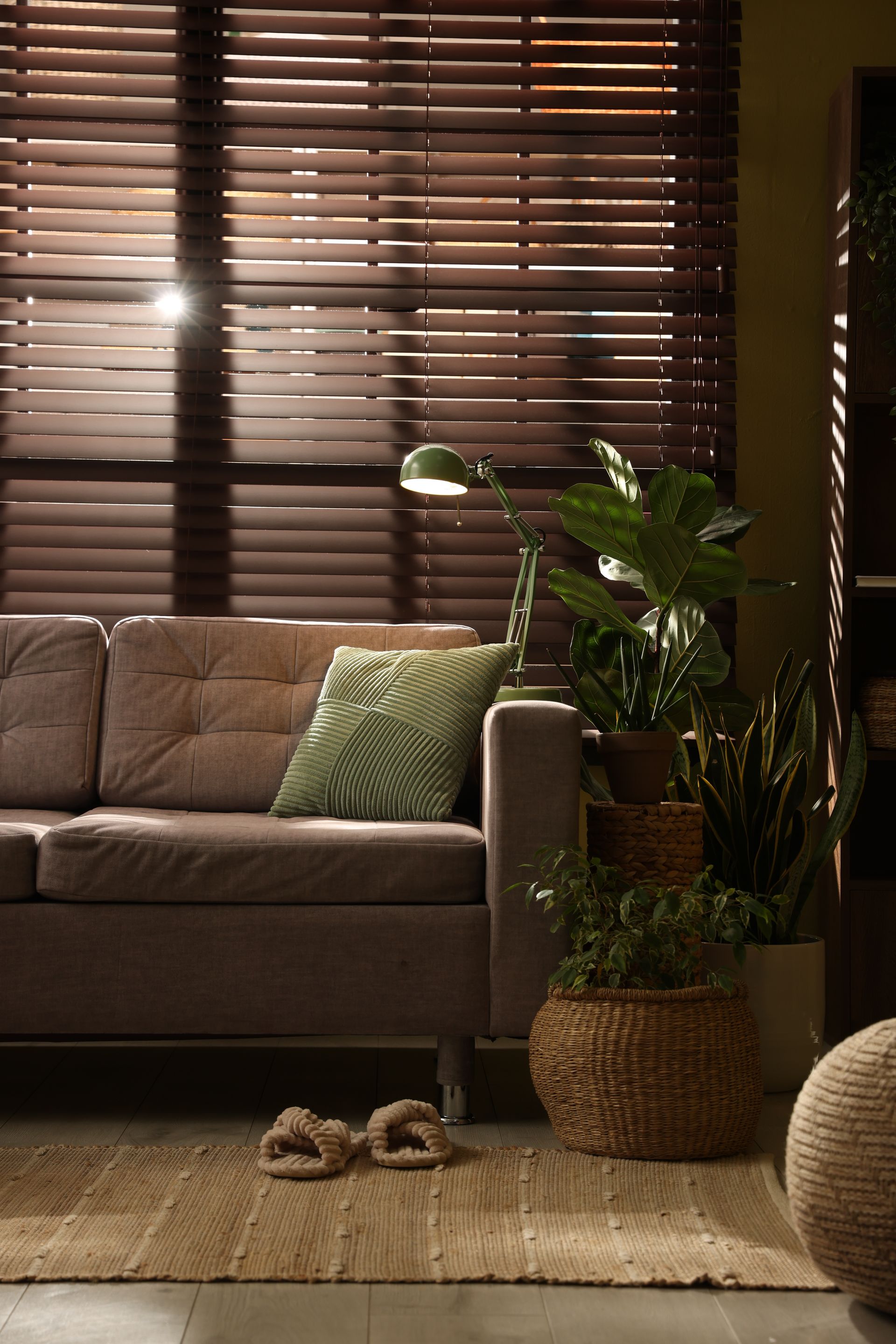 Cozy living room with gray sofa, green pillow, potted plant, woven rug, and window blinds in warm light