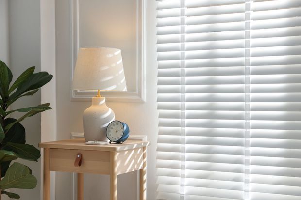 White lamp on a side table beside a plant and clock, next to bright window blinds