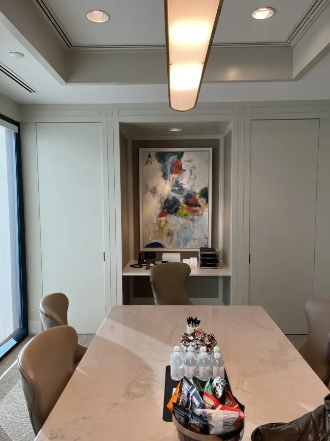 A conference room with a long table, chairs, an abstract painting, and snacks, lit by a rectangular ceiling fixture.