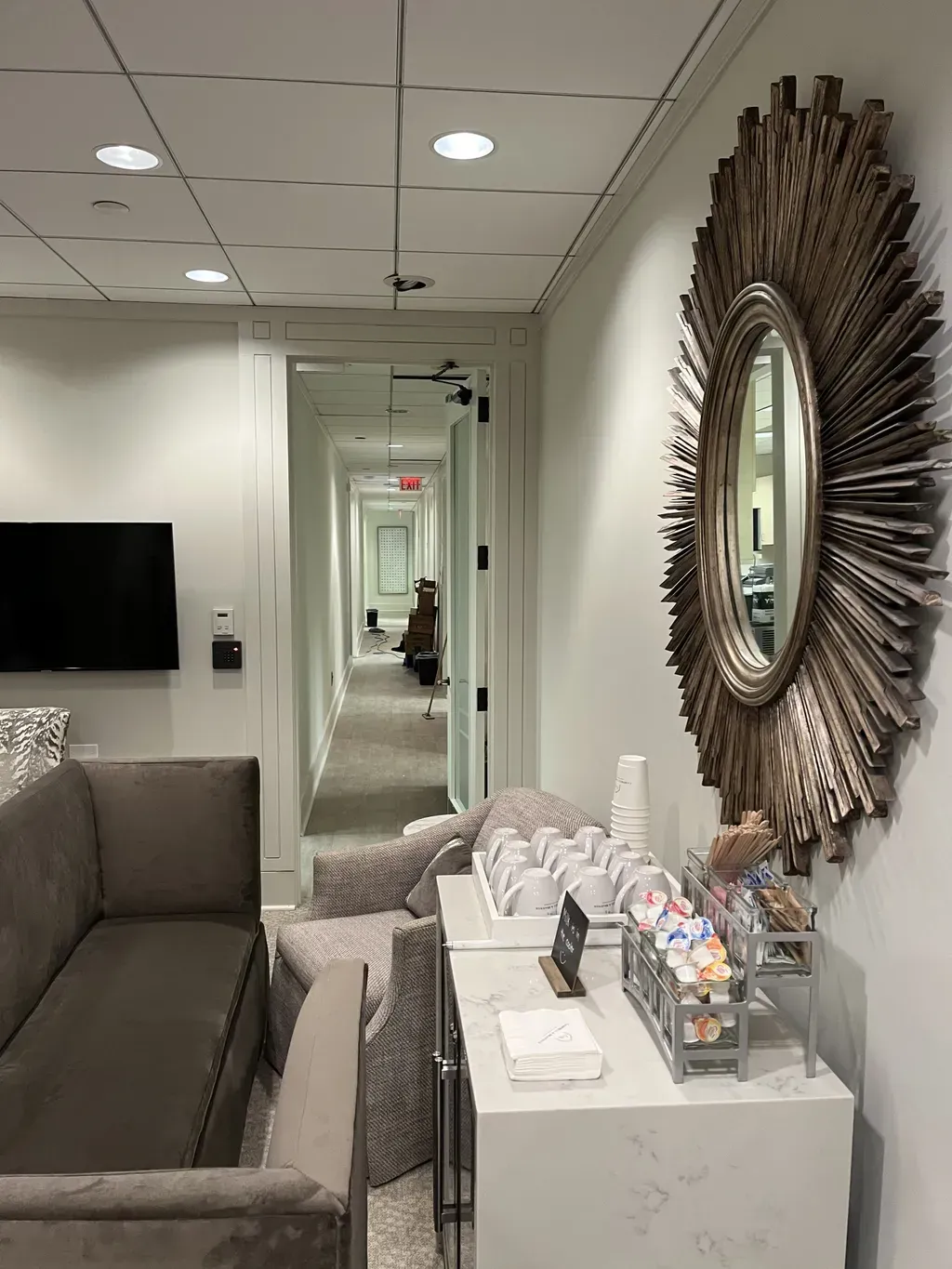 A neutral-toned waiting area with a sofa, a beverage station on a marble counter, and a large sunburst mirror on the wall.