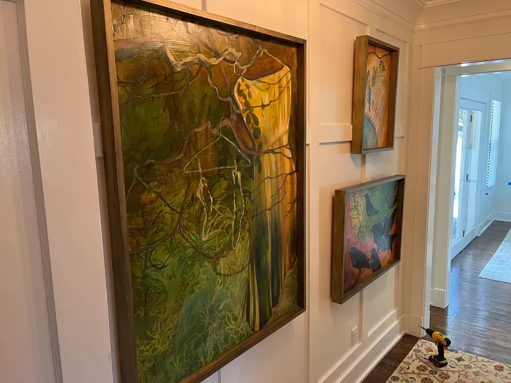 Three framed abstract paintings hang on a white paneled hallway wall, next to a doorway leading into a bright room.