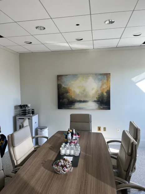 A conference room with a long wood table, four tan chairs, and an abstract landscape painting on a neutral wall.