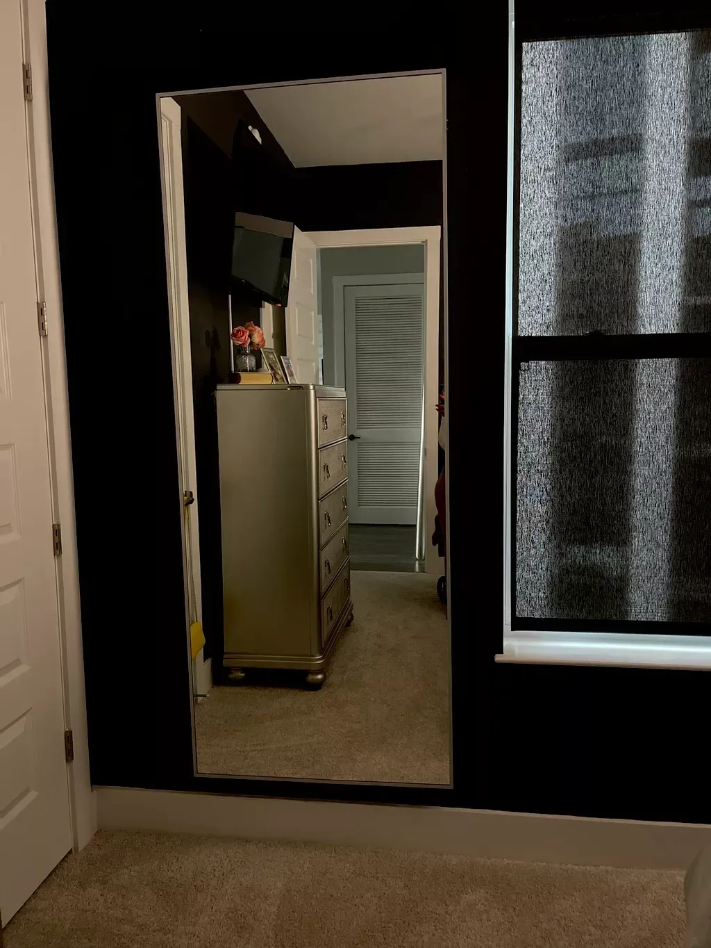 A floor-length mirror on a black wall reflects a room with a metallic dresser and an open doorway.