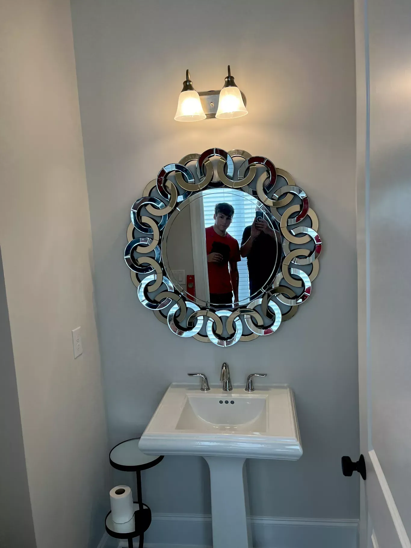A bathroom featuring a pedestal sink, a decorative circular mirror, and a wall-mounted light fixture on a gray wall.