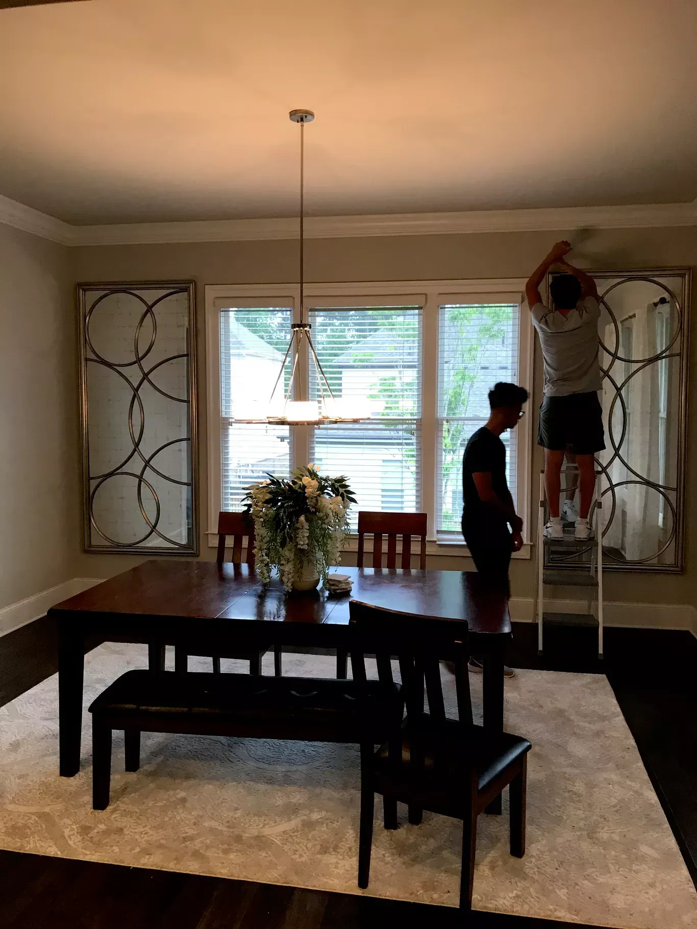 Two people install decorative wall mirrors in a dining room featuring a dark wood table, chairs, and a light-colored rug.