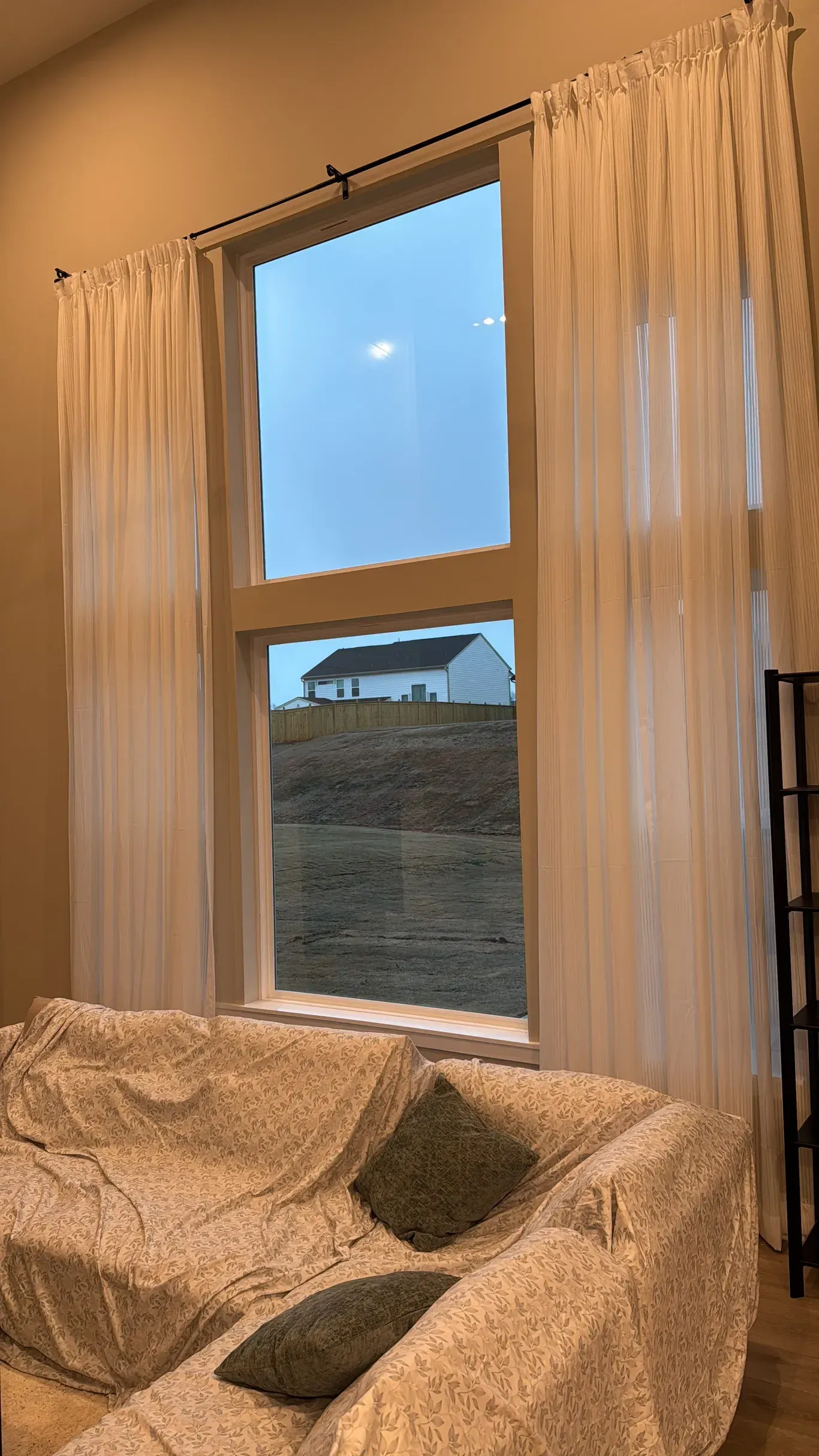 Tall white curtains flank a large two-story window looking out onto a rural landscape with a distant house.