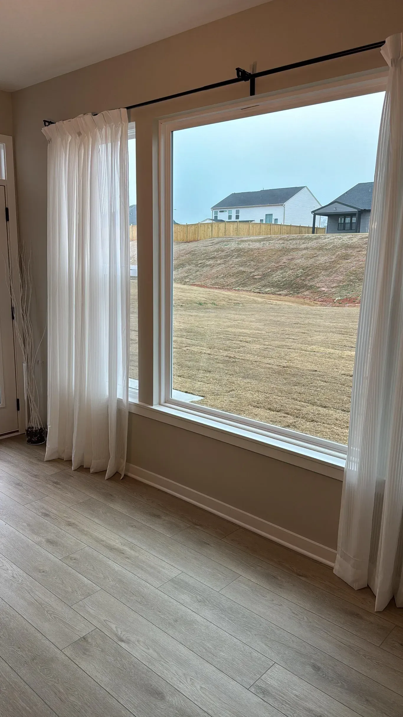A large window with sheer white curtains overlooks a backyard with a grassy slope and nearby houses under a cloudy sky.