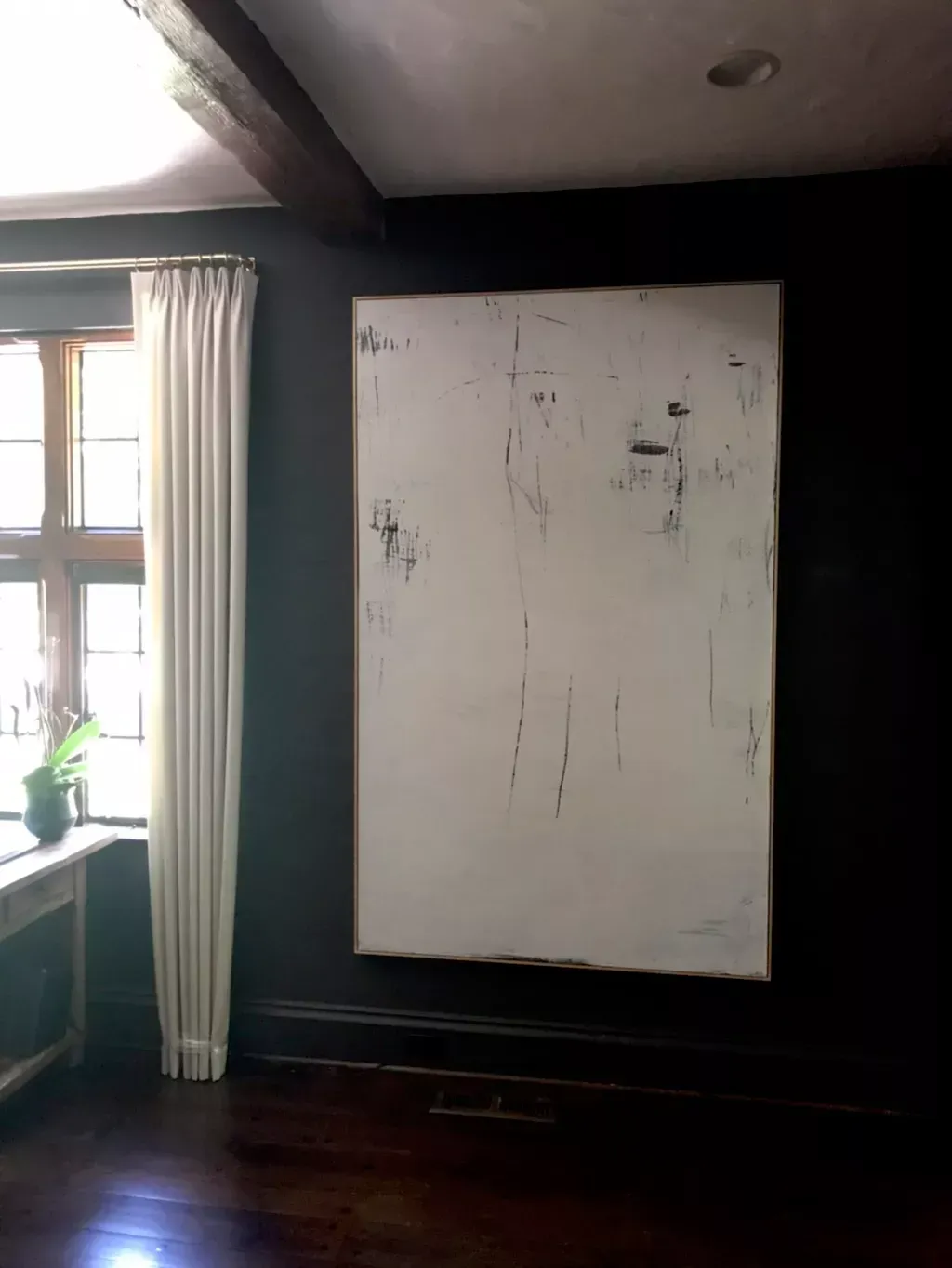 A large, abstract white painting with thin black markings hangs on a dark-walled room next to a window with white curtains.