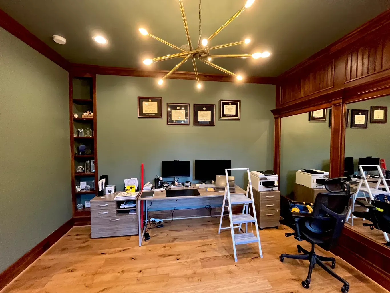 A home office with green walls, dark wood trim, a central desk, and a large mirror reflecting the space.