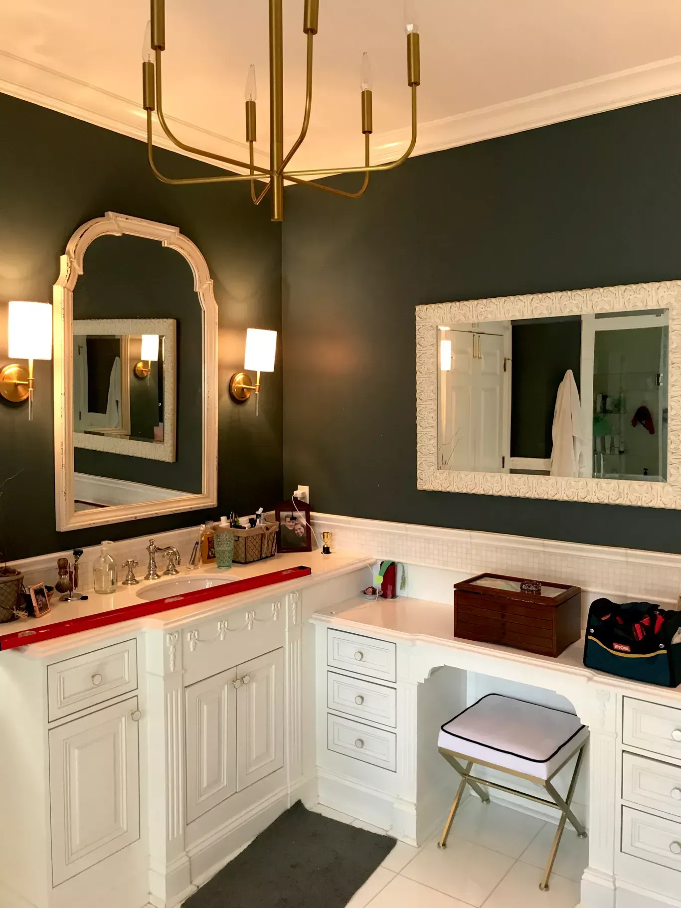 A bathroom with dark green walls, white cabinets, a brass chandelier, two mirrors, and a vanity stool.