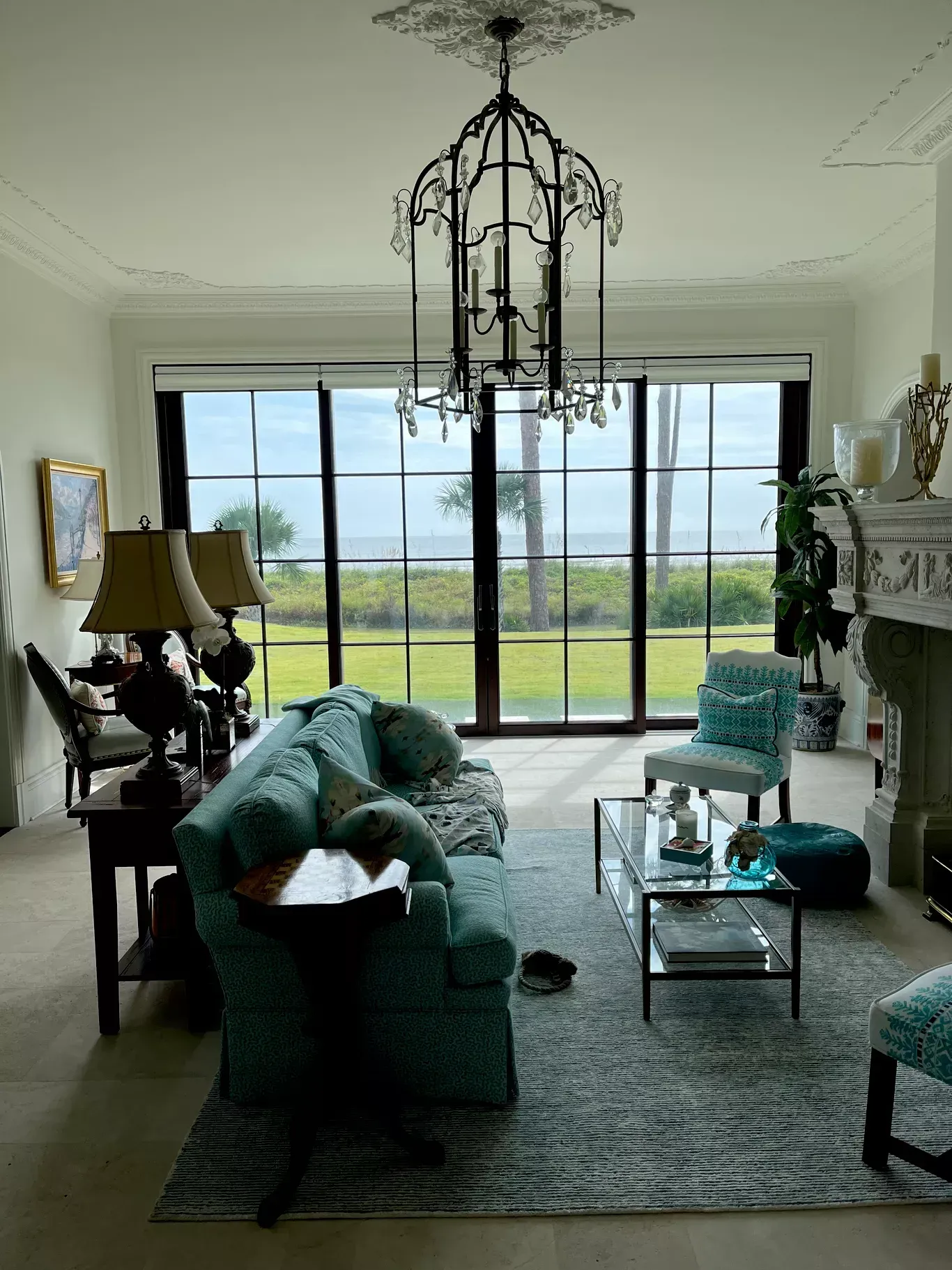 A living room with a teal sofa, glass coffee table, and large window wall overlooking a green lawn and trees.