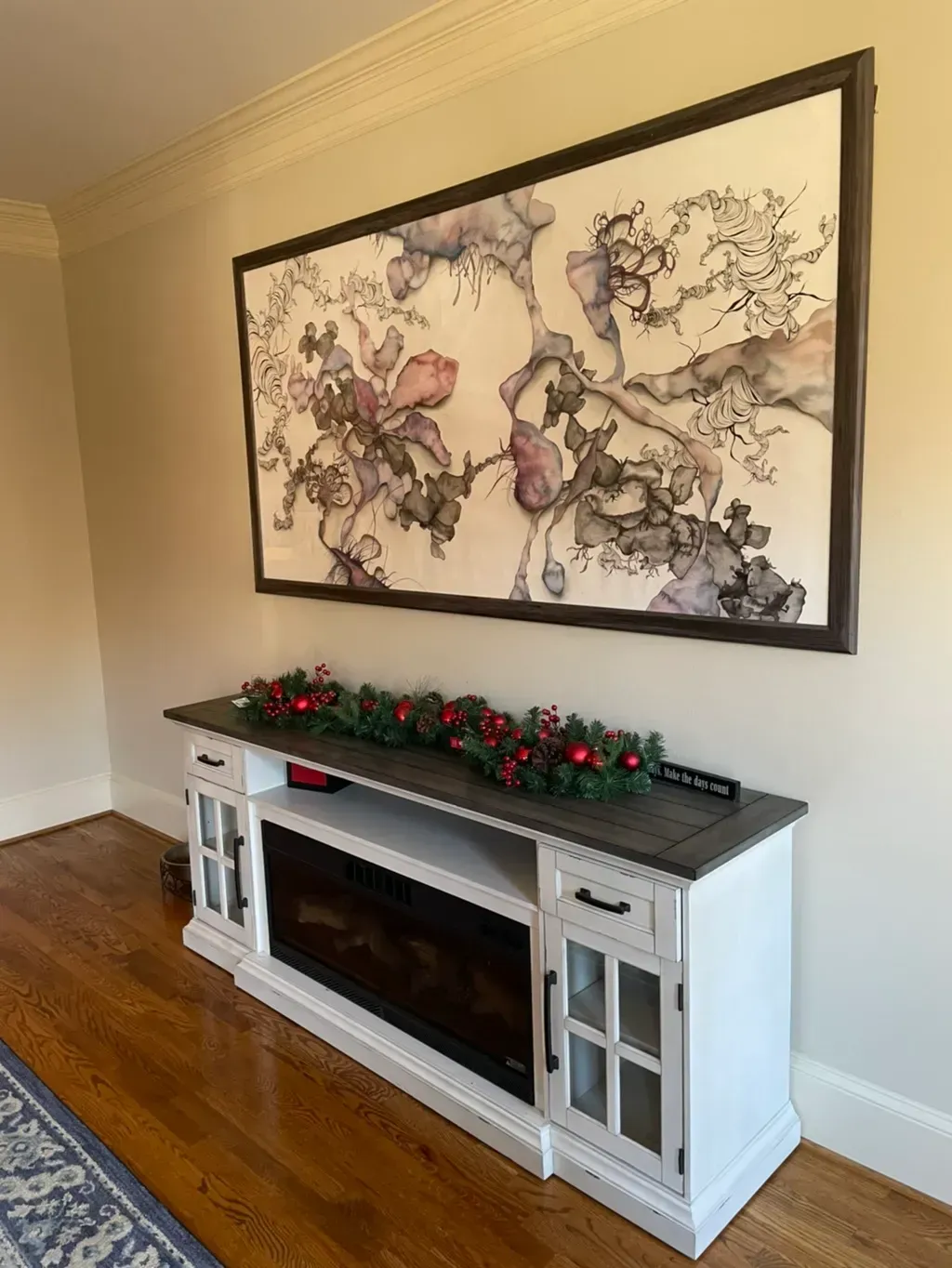 A white fireplace console with a dark wood top and holiday greenery sits beneath a large, framed botanical art piece.