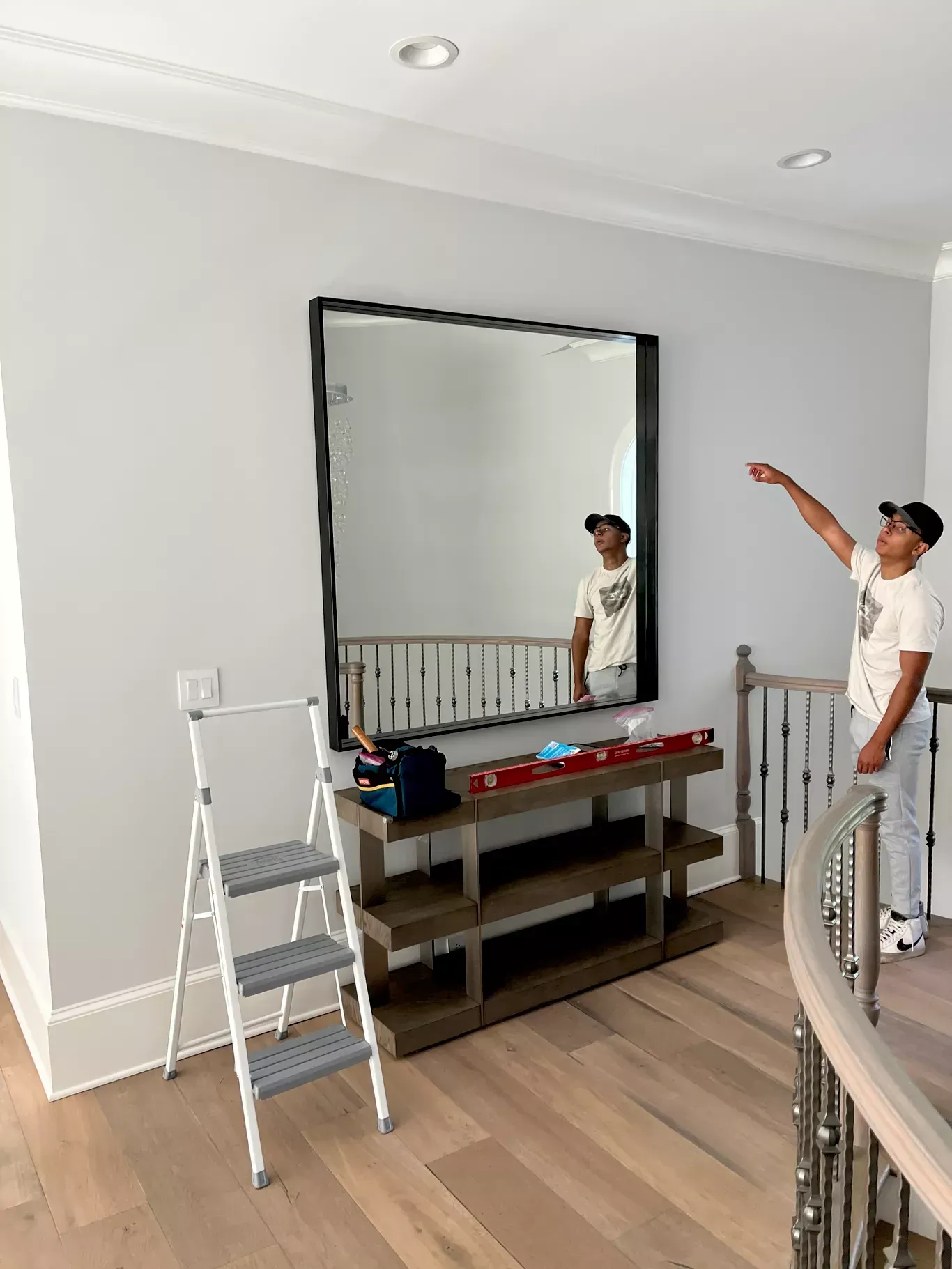 A person in a white t-shirt points toward a large square mirror mounted on a light gray wall above a tiered wooden console.