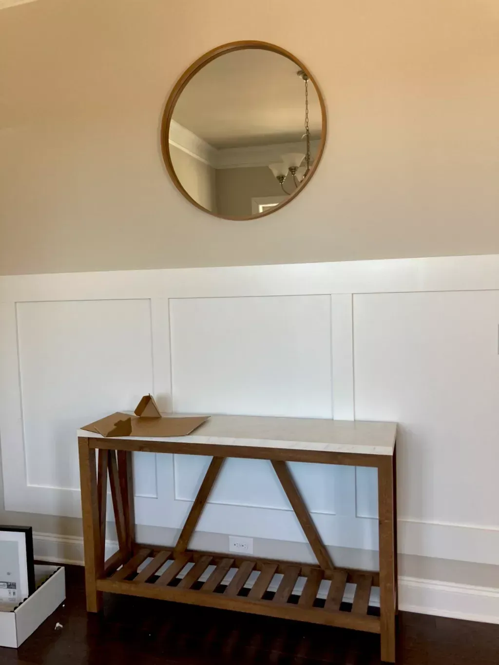 A round wall mirror hangs above a wooden console table with a white top against a wall with white wainscoting.