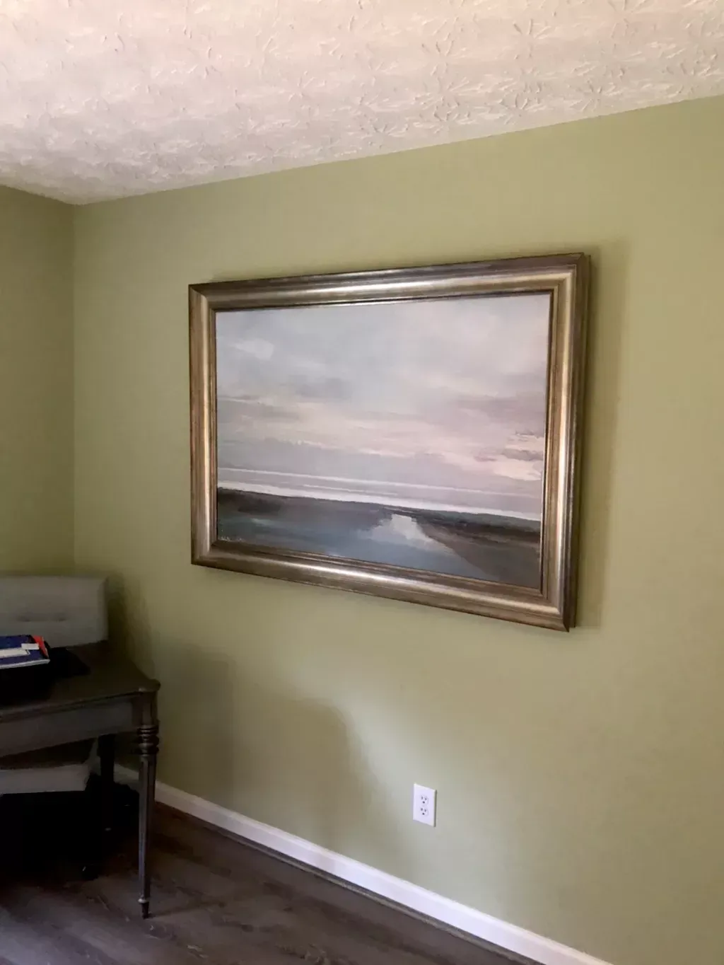 A landscape painting in a metallic frame hangs on a sage green wall above a desk and an electrical outlet.