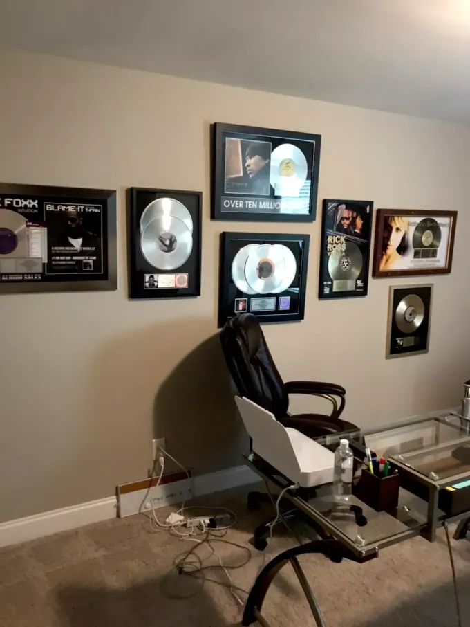 A collection of framed gold and platinum record awards mounted on a beige wall above a home office desk and chair.