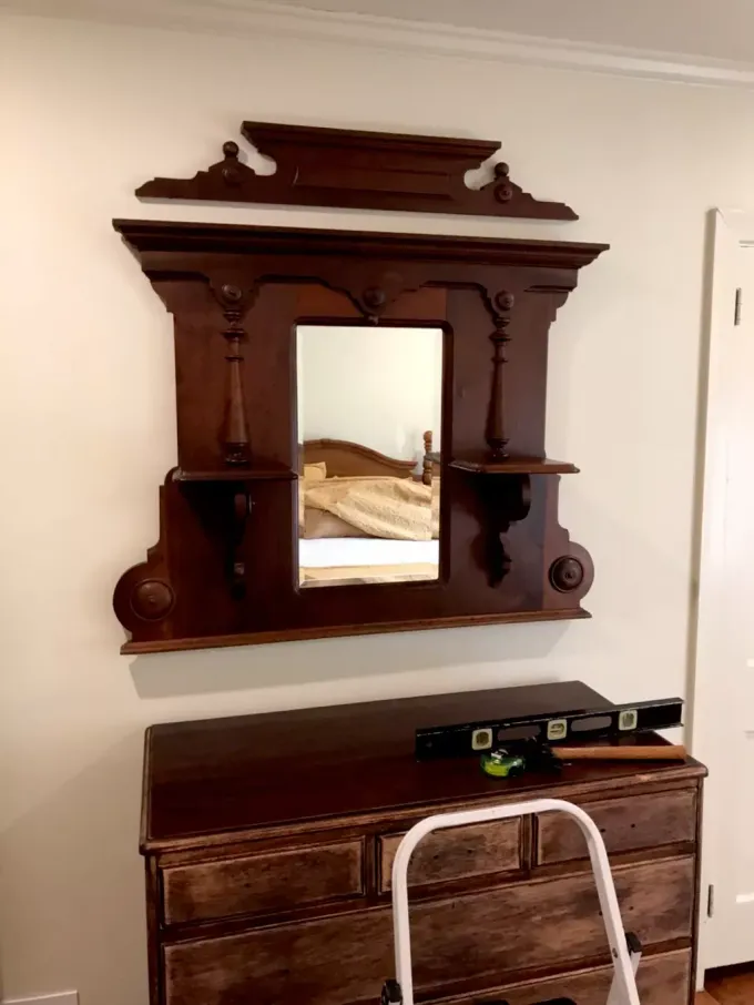 A dark wood Victorian-style mirror frame hanging above a wooden dresser, with a loose decorative crest piece above it.