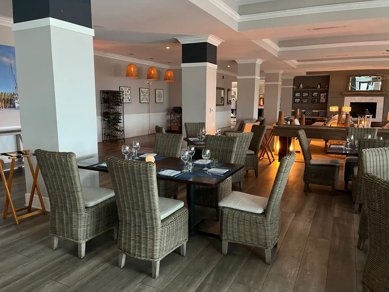 A modern restaurant dining room featuring woven chairs, dark tables, light gray walls, and pendant lighting.