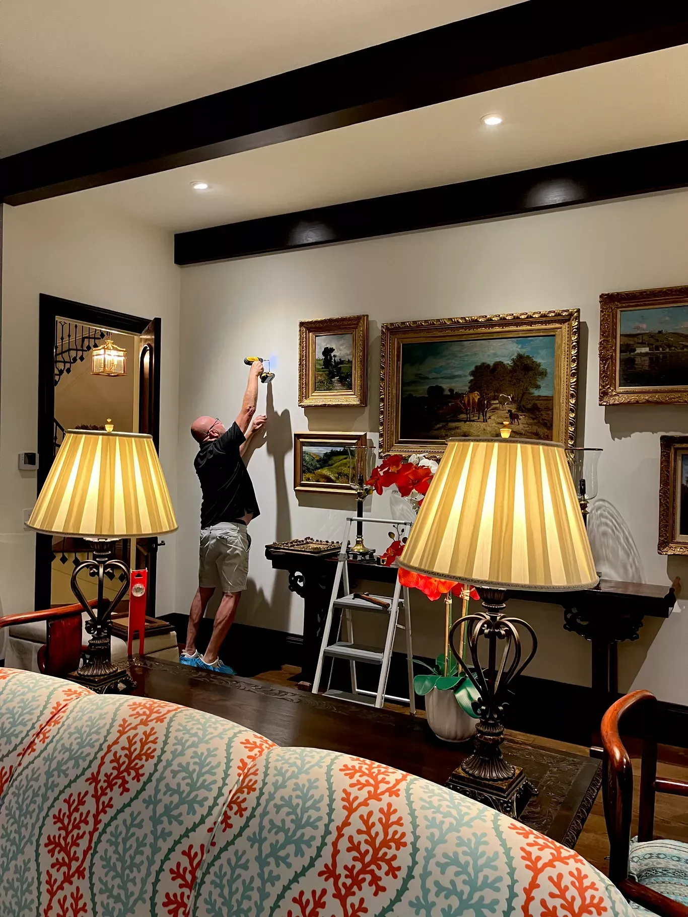 A person hangs art on a gallery wall near lamps in a living room, with a stepladder nearby.
