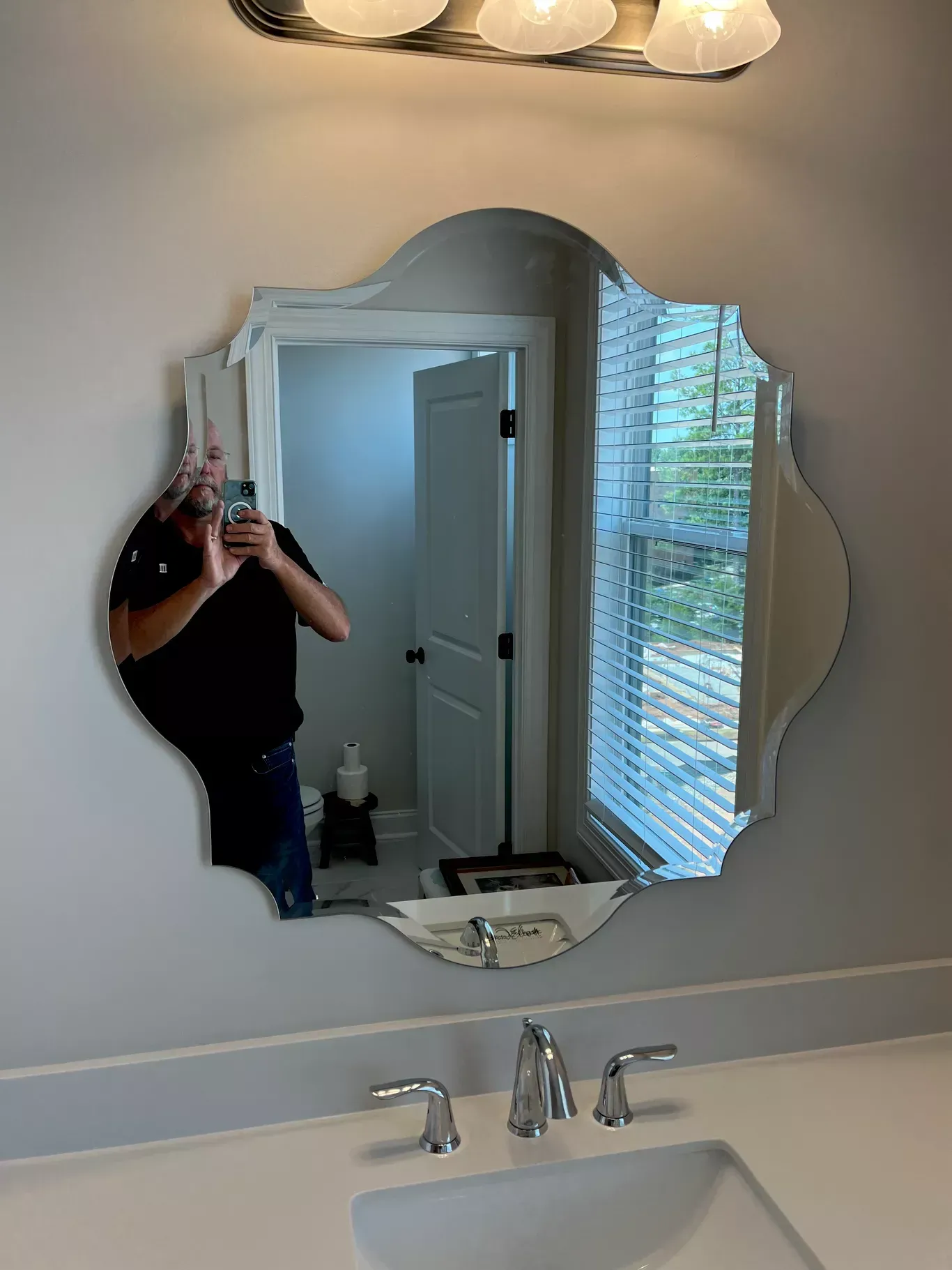 A scalloped bathroom mirror reflects a person holding a phone, mounted above a sink and faucet against a plain wall.