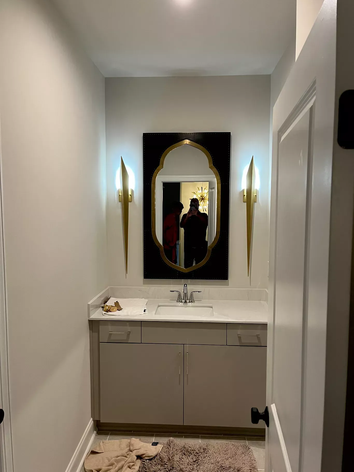A bathroom vanity with a black-framed ornate mirror, gold wall sconces, and neutral cabinetry, seen through a doorway.