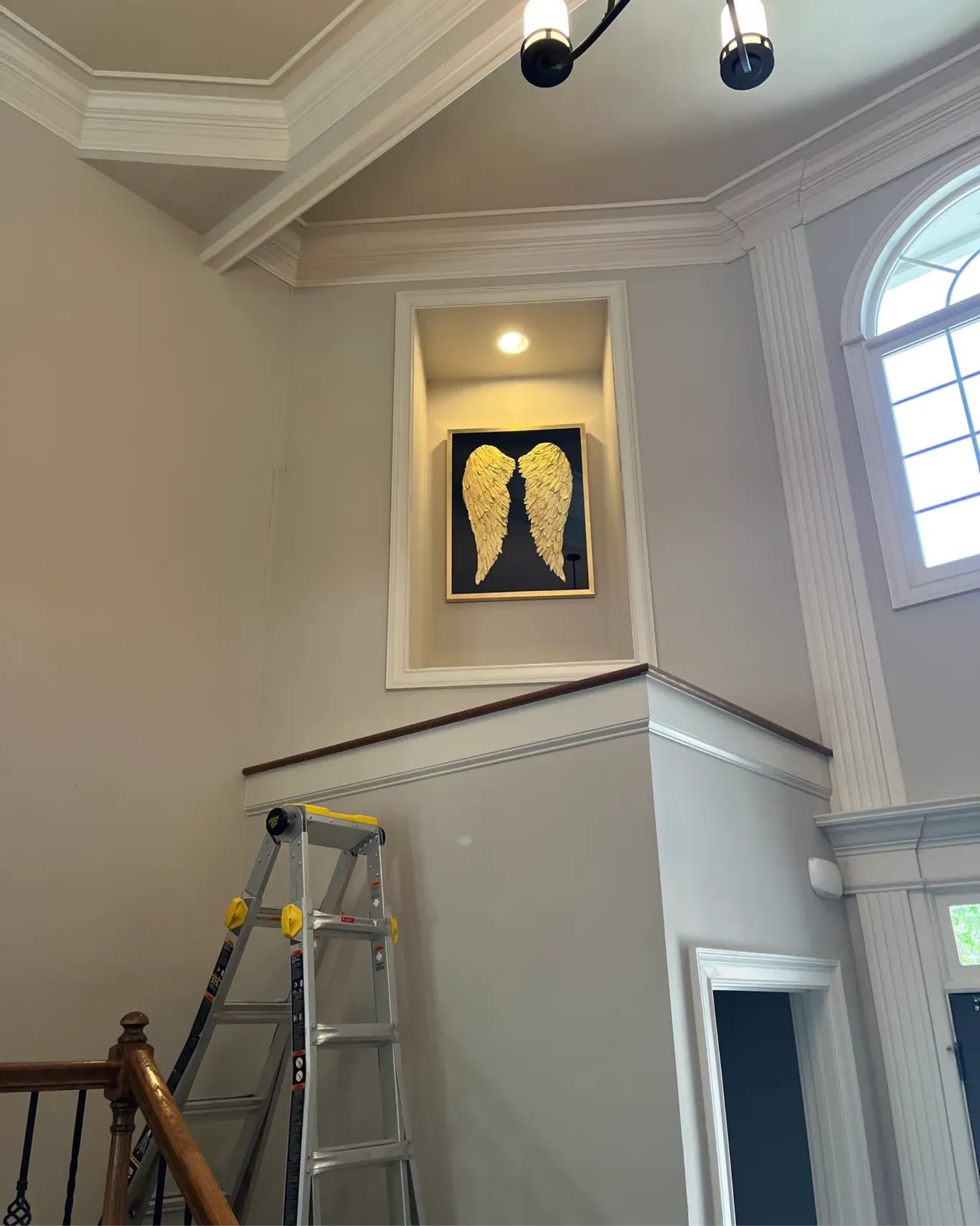 An indoor shot of a tall wall with a recessed alcove holding gold angel wing art above a window and stair railing.