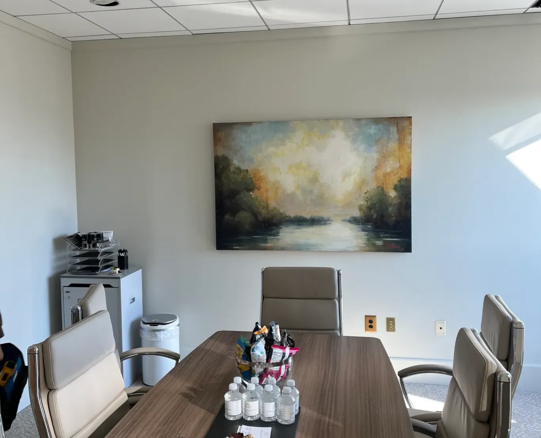 A conference room with a wood table, rolling chairs, and an abstract landscape painting hanging on a light gray wall.