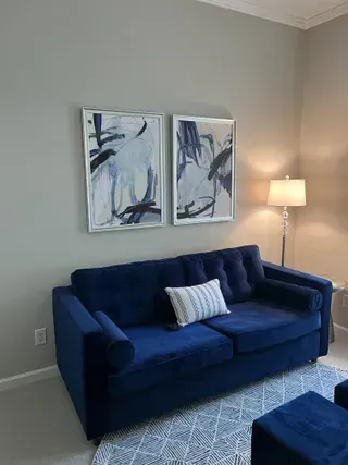 A navy blue tufted sofa with a patterned pillow, sits below two abstract blue-toned paintings and next to a floor lamp.