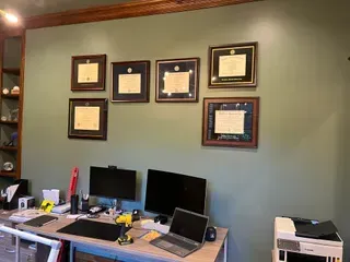 A home office with a desk featuring two monitors and a laptop, with six framed diplomas displayed on a sage green wall.