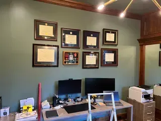 A home office desk sits against a sage green wall adorned with a gallery of seven framed degrees and a military medal set.