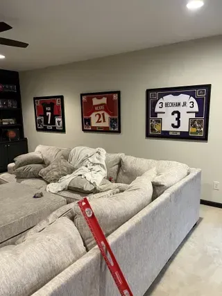 Three framed sports jerseys hang on a light wall above a large, beige sectional sofa, with a level held in the foreground.