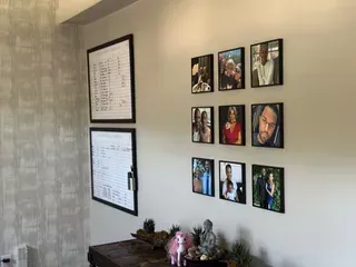 A wall features two framed documents and a grid of nine square photos above a wooden table with small decorative items.