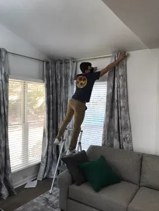 A person standing on a step stool on a couch, reaching up to adjust a curtain rod above a window.